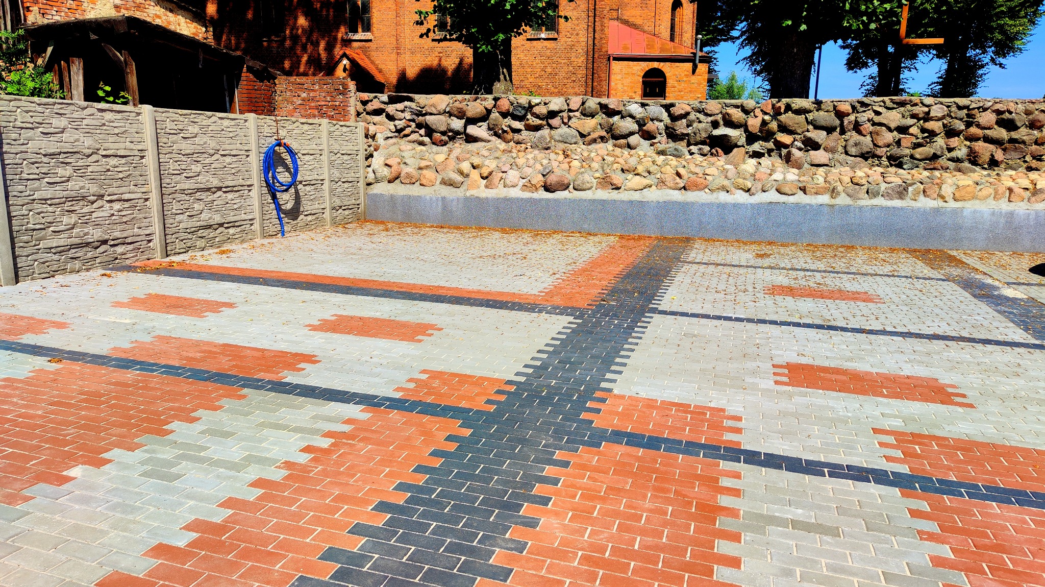 Wzór z kostki brukowej w odcieniach czerwieni, szarości i czerni tworzący geometryczny design na podwórku, z widocznym ogrodzeniem betonowym o fakturze kamienia i fragmentem budynku w tle.