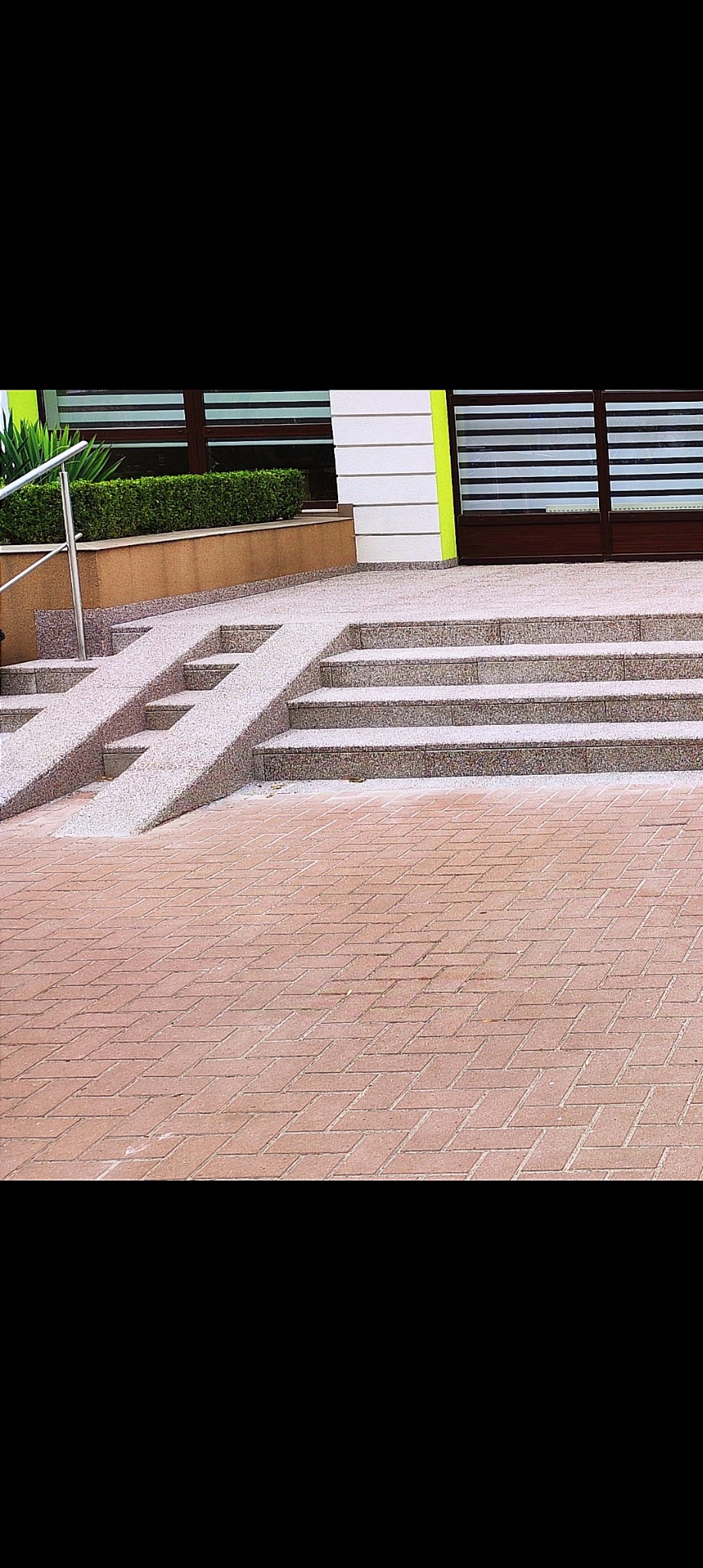 Wejście do budynku z rampą dla niepełnosprawnych i schodami, wykończone kostką brukową w ceglanym odcieniu, z elementami zieleni w tle i nowoczesną fasadą.