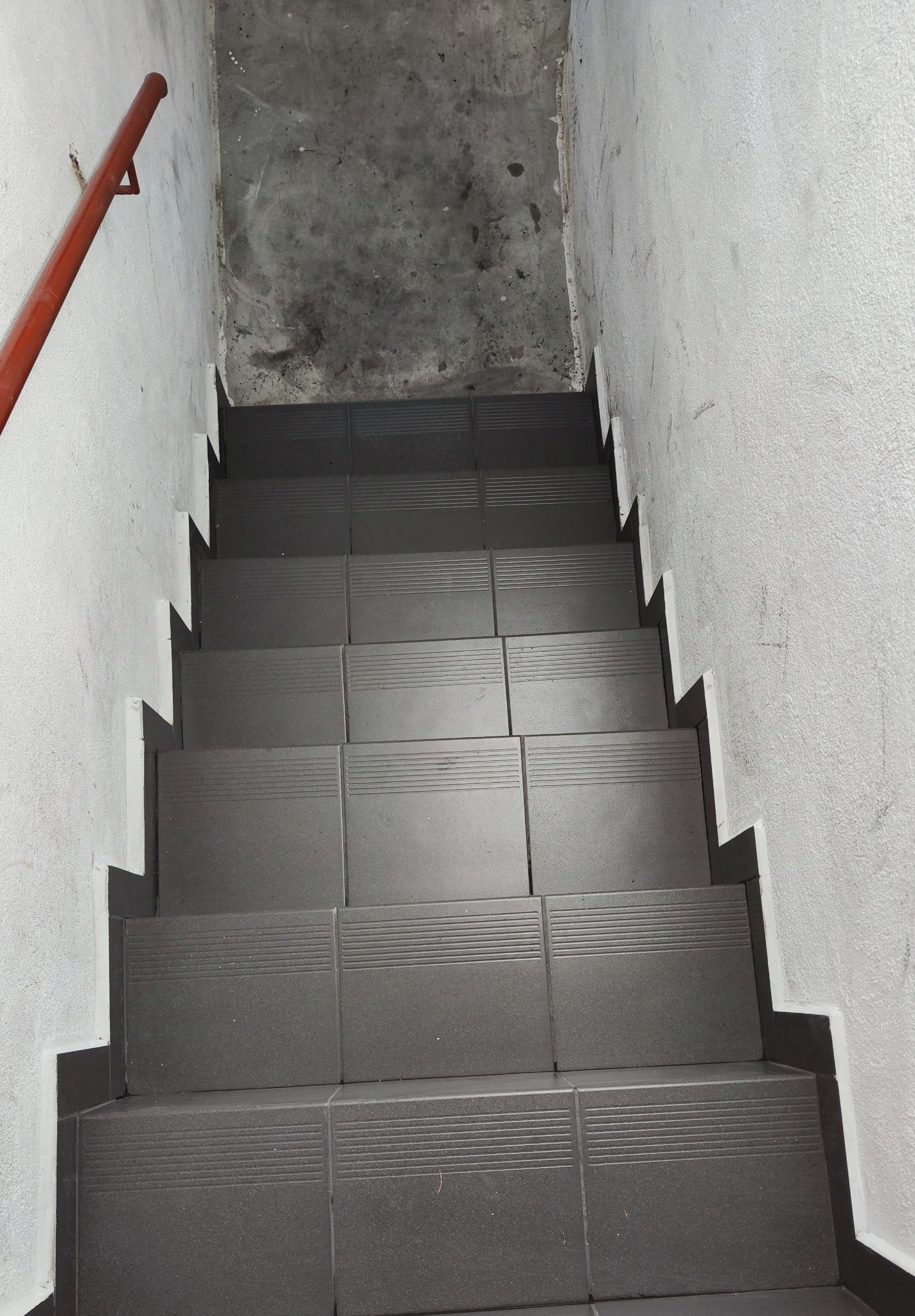 Schody wyłożone szarymi płytkami z poziomymi rowkami antypoślizgowymi, widok z góry, czerwona poręcz po lewej stronie, ściany w odcieniach bieli i szarości.