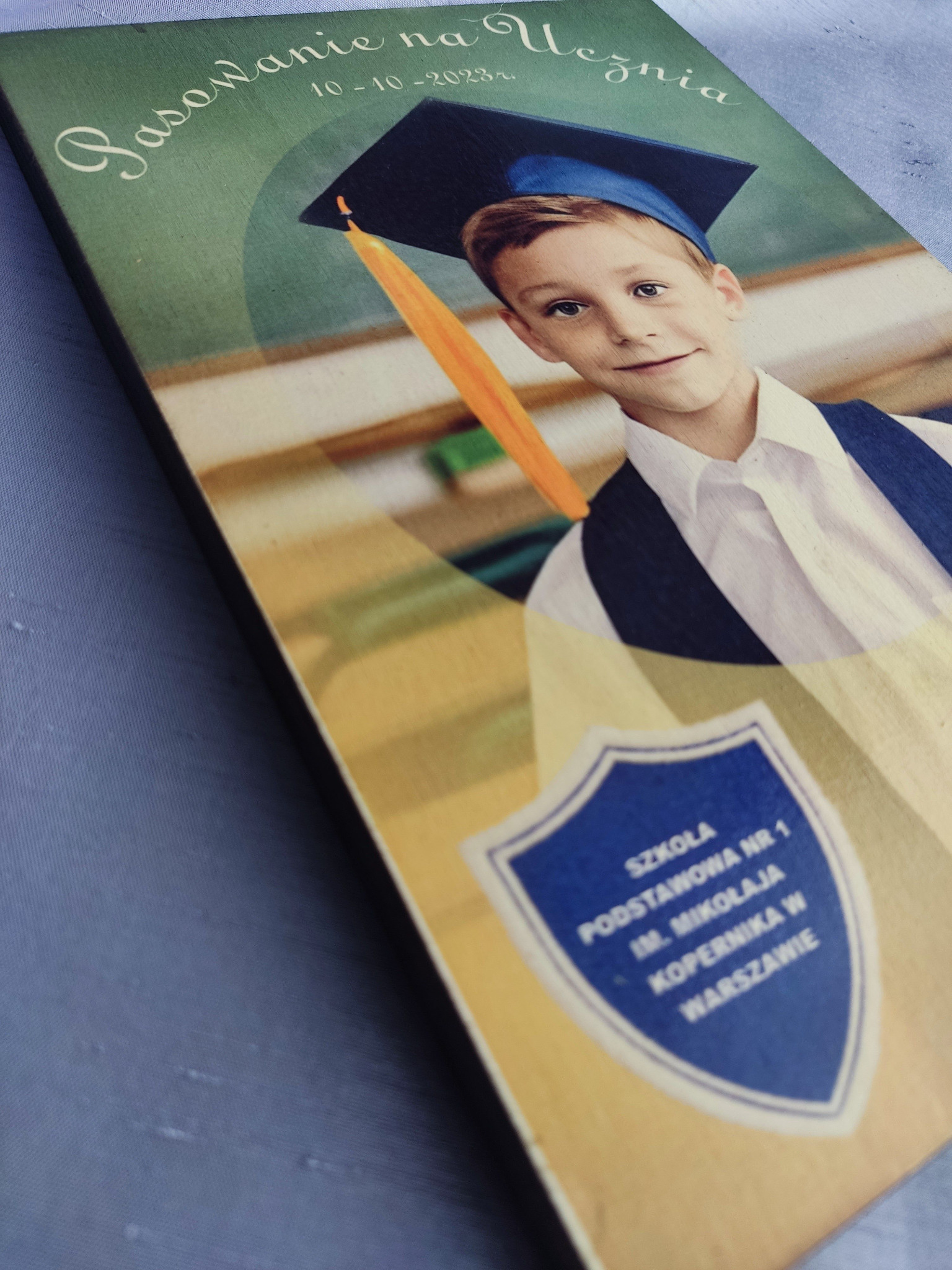 Pamiątkowa tablica z wizerunkiem chłopca w todze i birecie z okazji pasowania na ucznia, z datą 10-10-2023 i nazwą Szkoły Podstawowej nr 1 im. Mikołaja Kopernika w Warszawie.