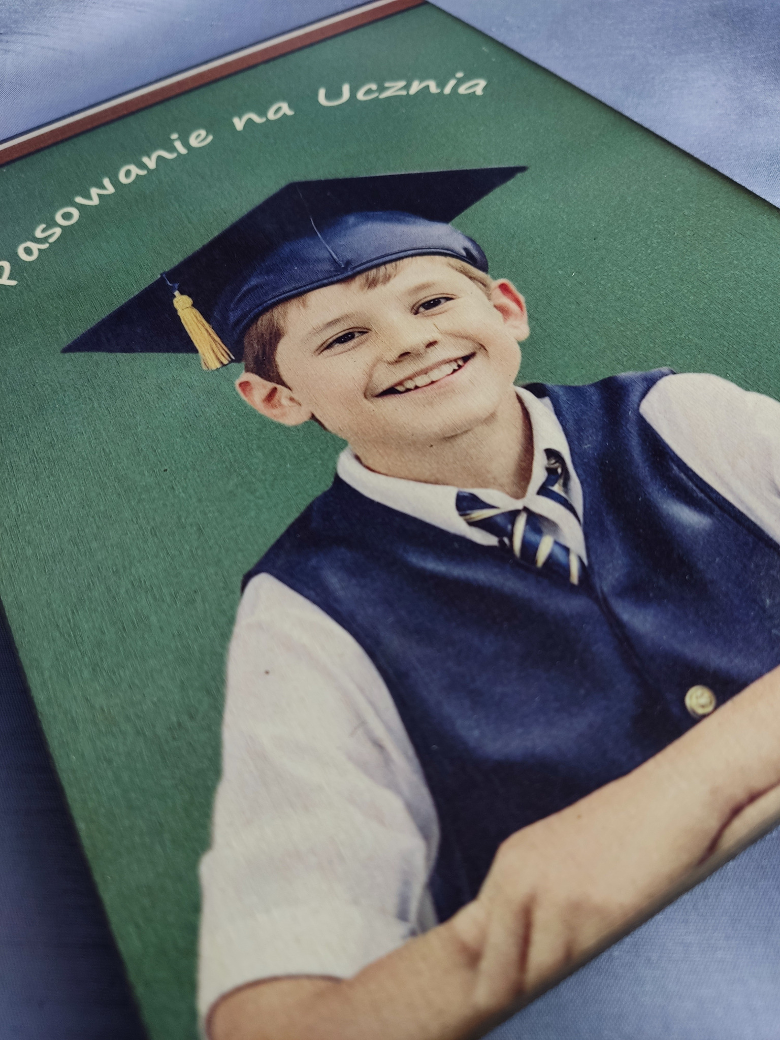 Portret uśmiechniętego chłopca w todze absolwenta na tle zielonej tablicy z napisem 'Pasowanie na Ucznia', zdjęcie w formacie zbliżonym do polaroidu.