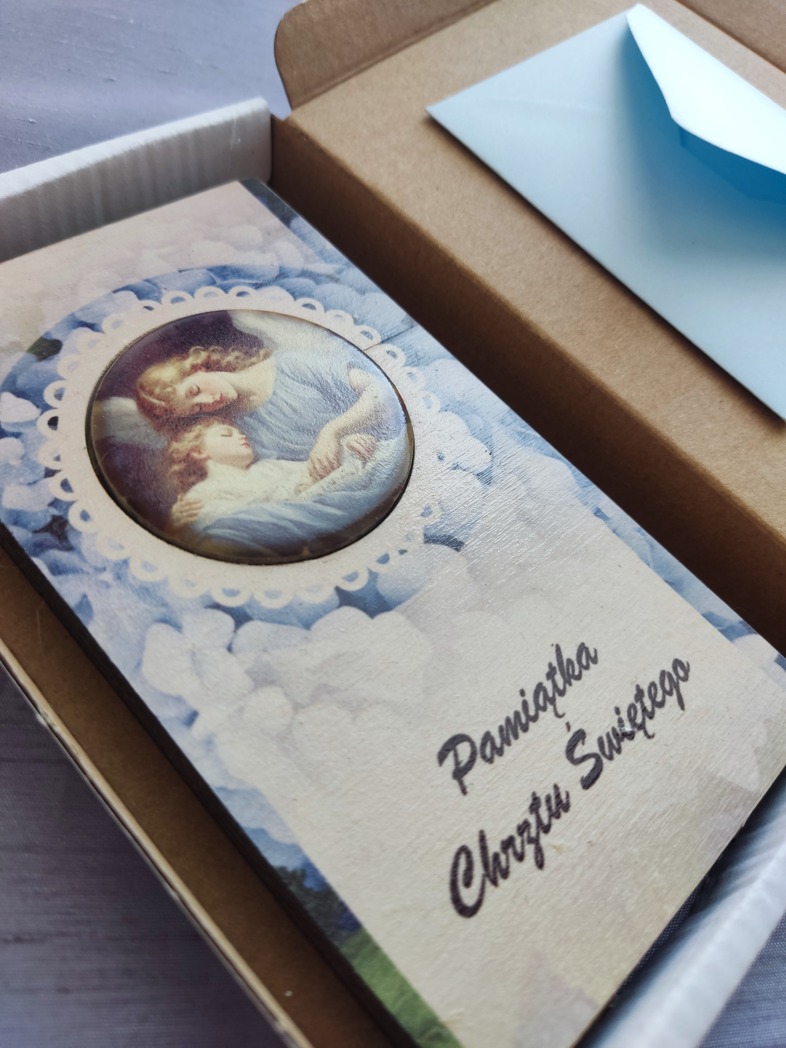 Drewniana pamiątka Chrztu Świętego z wizerunkiem Matki Boskiej z Dzieciątkiem, umieszczona w kartonowym pudełku obok błękitnej koperty.