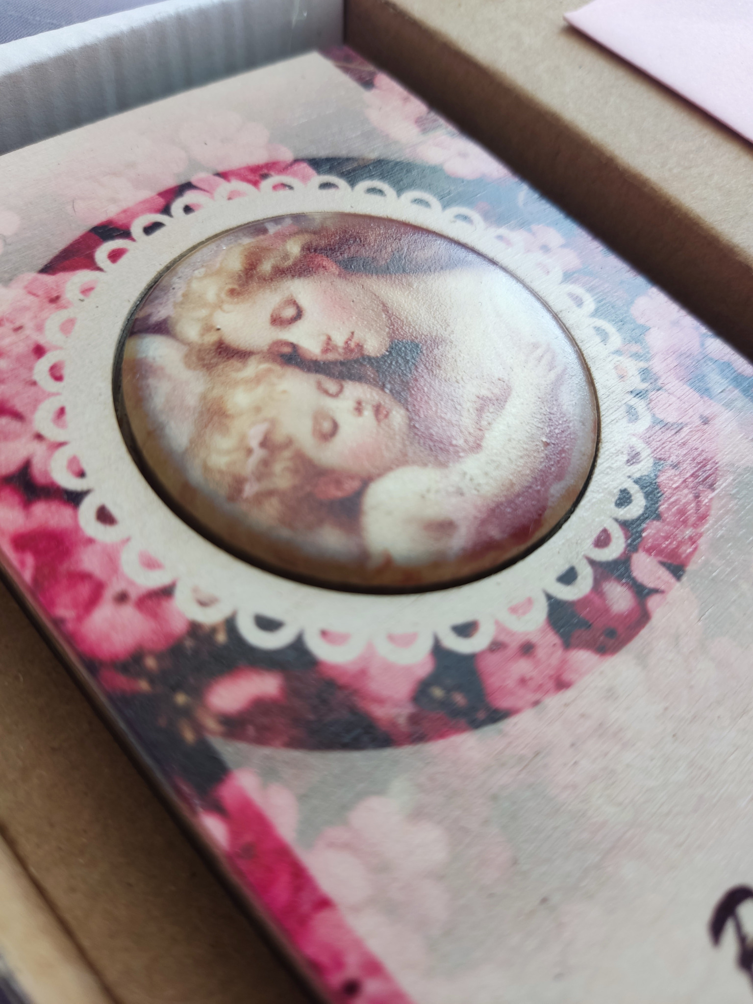 Drewniane pudełko z okrągłą, ozdobną ramką i portretem dziecka w sepii, otoczone motywem różowych kwiatów.