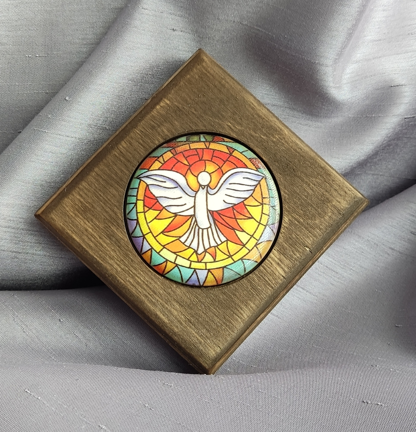 Drewniana ramka w kształcie kwadratu z okrągłym obrazkiem gołębicy w stylu witrażu na tle szarego materiału.
