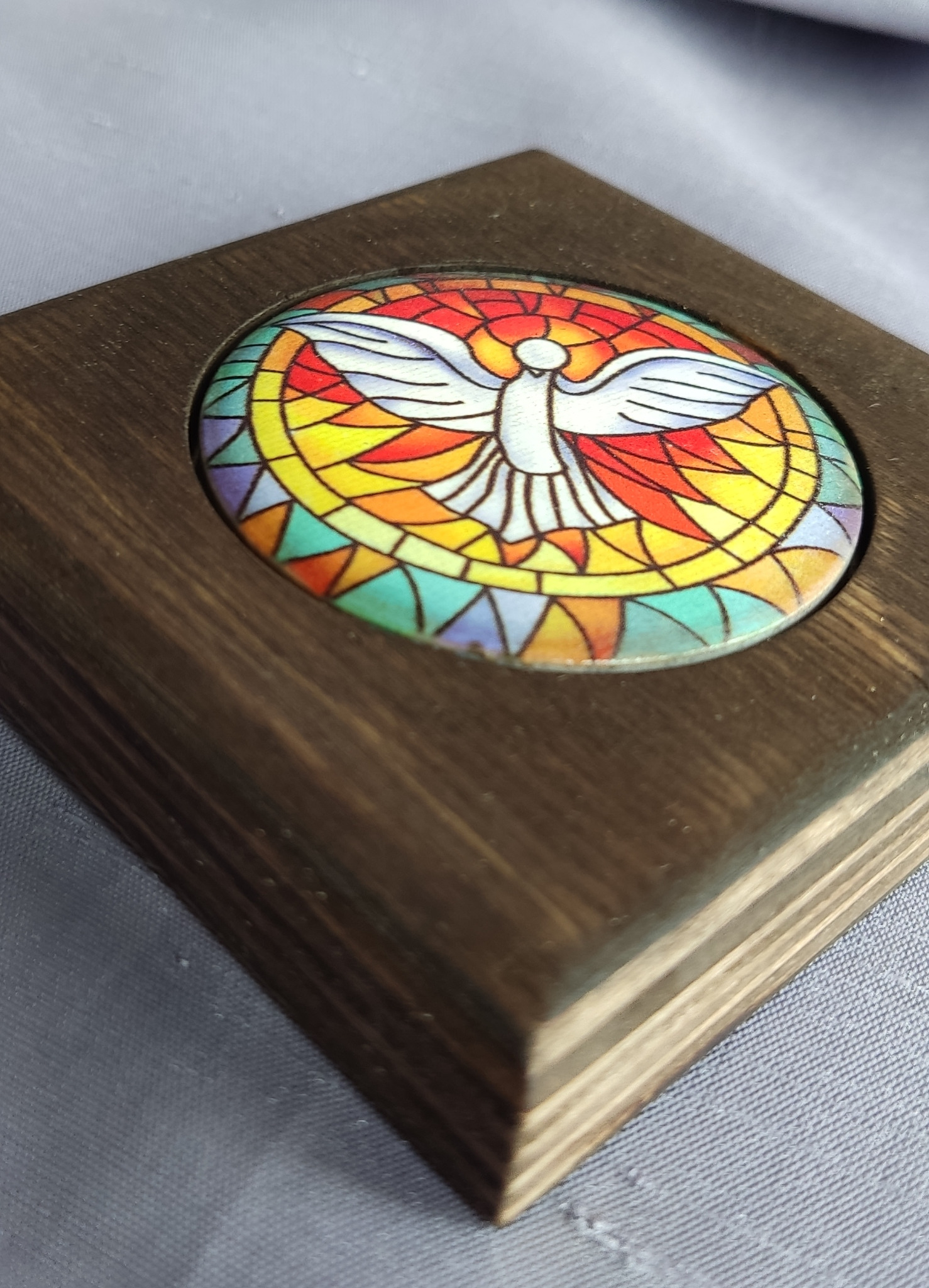 Drewniana ramka z kolorowym, okrągłym witrażem przedstawiającym gołębicę, symbol Ducha Świętego, na szarym tle.