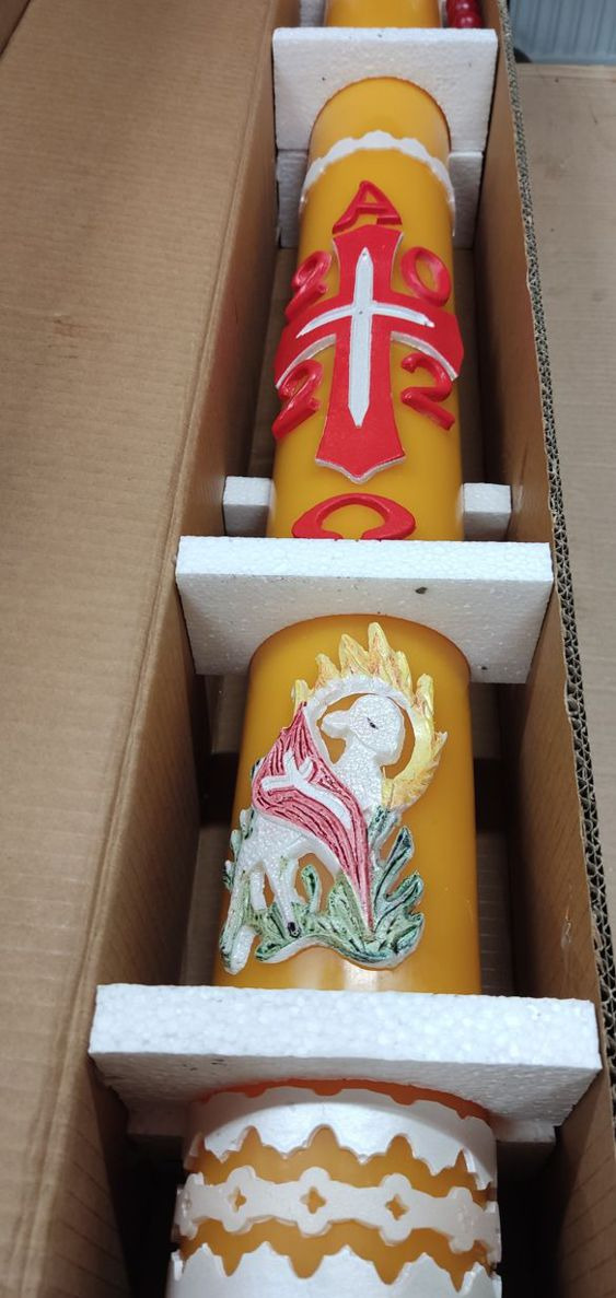 Dwie zdobione świece w odcieniach żółci i bieli, zabezpieczone styropianem wewnątrz kartonowego pudełka. Górna świeca z czerwonym krzyżem i literami Alfa i Omega, dolna z motywem Baranka Bożego.