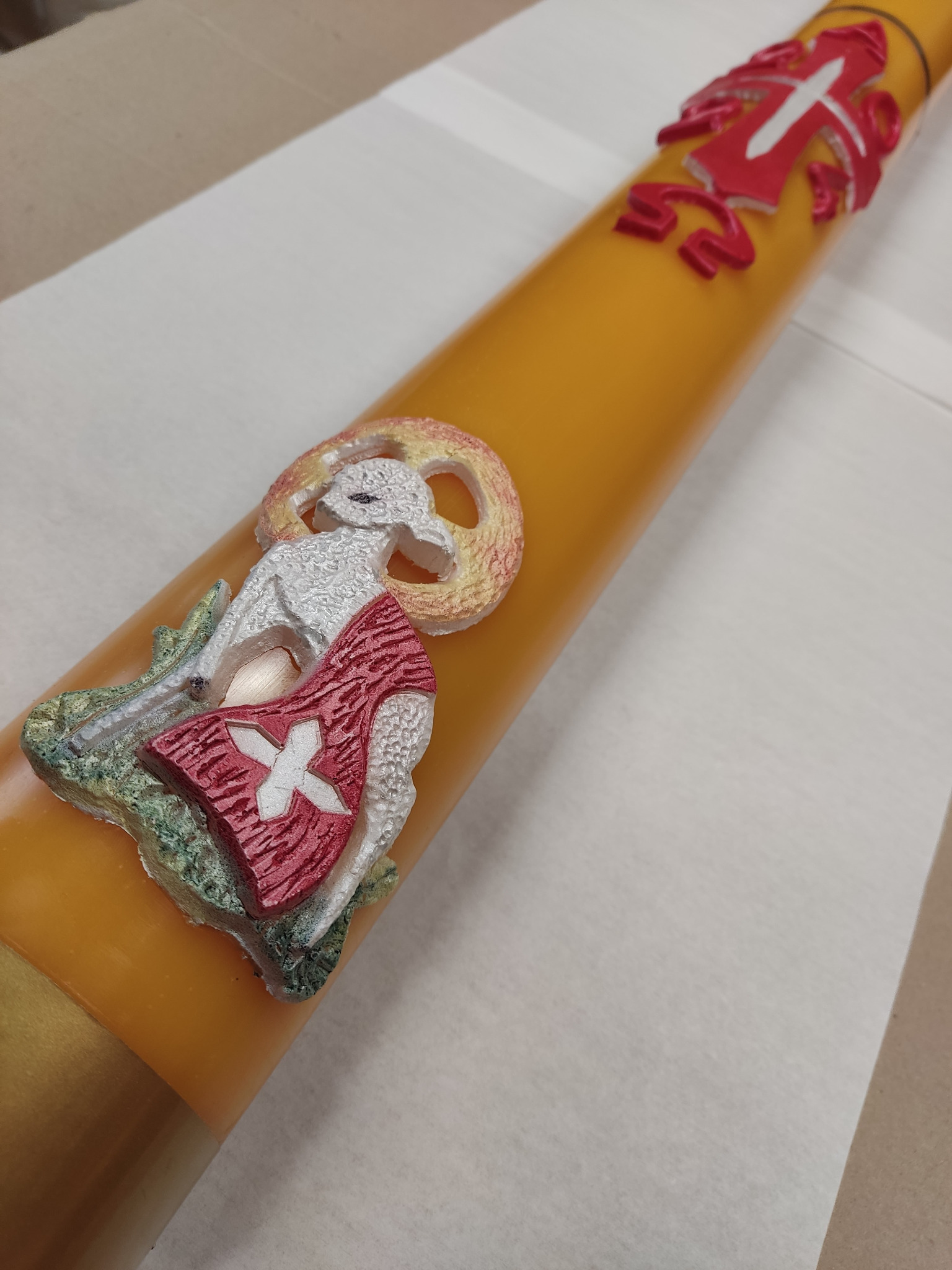 Żółta świeca paschalna z wizerunkiem Baranka Bożego z czerwoną szatą i białym krzyżem szwajcarskim, umieszczona na białym papierze.