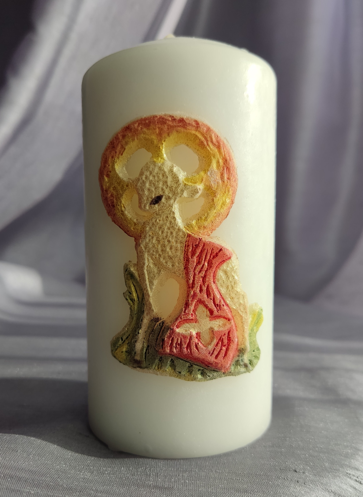 Biała świeca z wytłoczonym barankiem paschalnym w kolorach czerwieni, żółci i zieleni na tle delikatnej tkaniny.