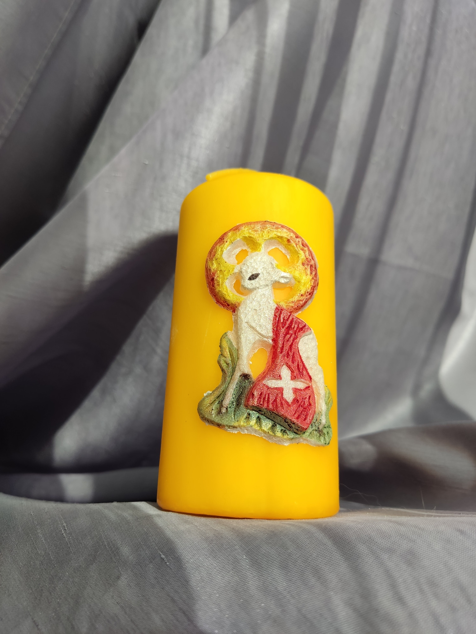 Żółta świeca z wizerunkiem baranka z aureolą i czerwonym okryciem z białym krzyżem, na tle szarej, drapowanej tkaniny.