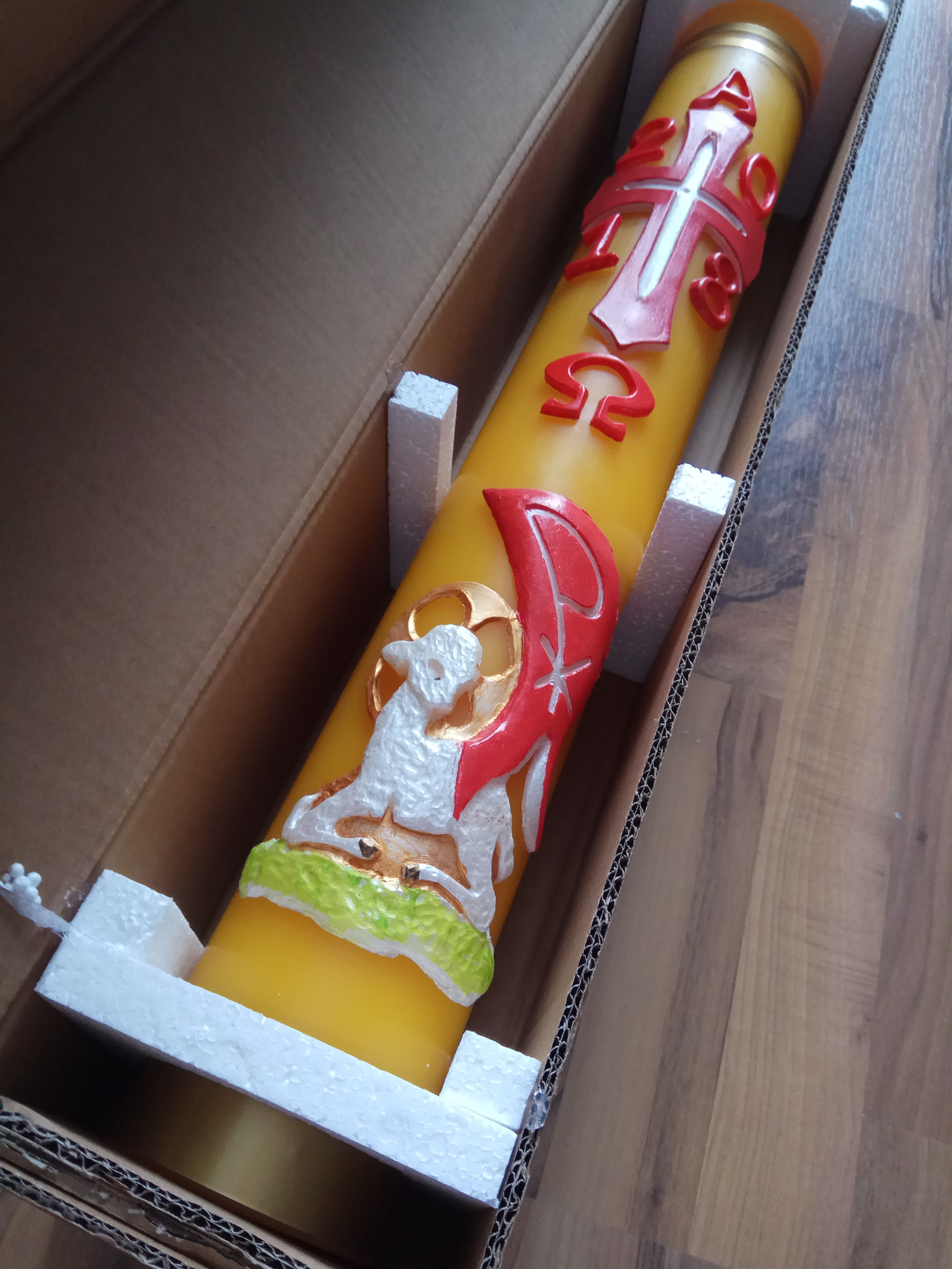 Żółta, zdobiona świeca paschalna z motywami religijnymi (krzyż, baranek) w kartonowym pudełku z piankowym zabezpieczeniem.