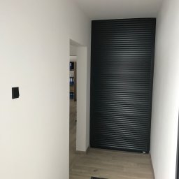 CHMUR-BUD - Wnętrze korytarza po remoncie z białymi ścianami, drewnopodobną podłogą i czarną roletą antywłamaniową zasłaniającą wejście, widoczny włącznik światła i oświetlenie punktowe.