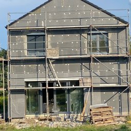 CHMUR-BUD - Dom w trakcie ocieplania styropianem grafitowym, widoczne rusztowania, okna z roletami zewnętrznymi, paleta drewniana leżąca na trawie.