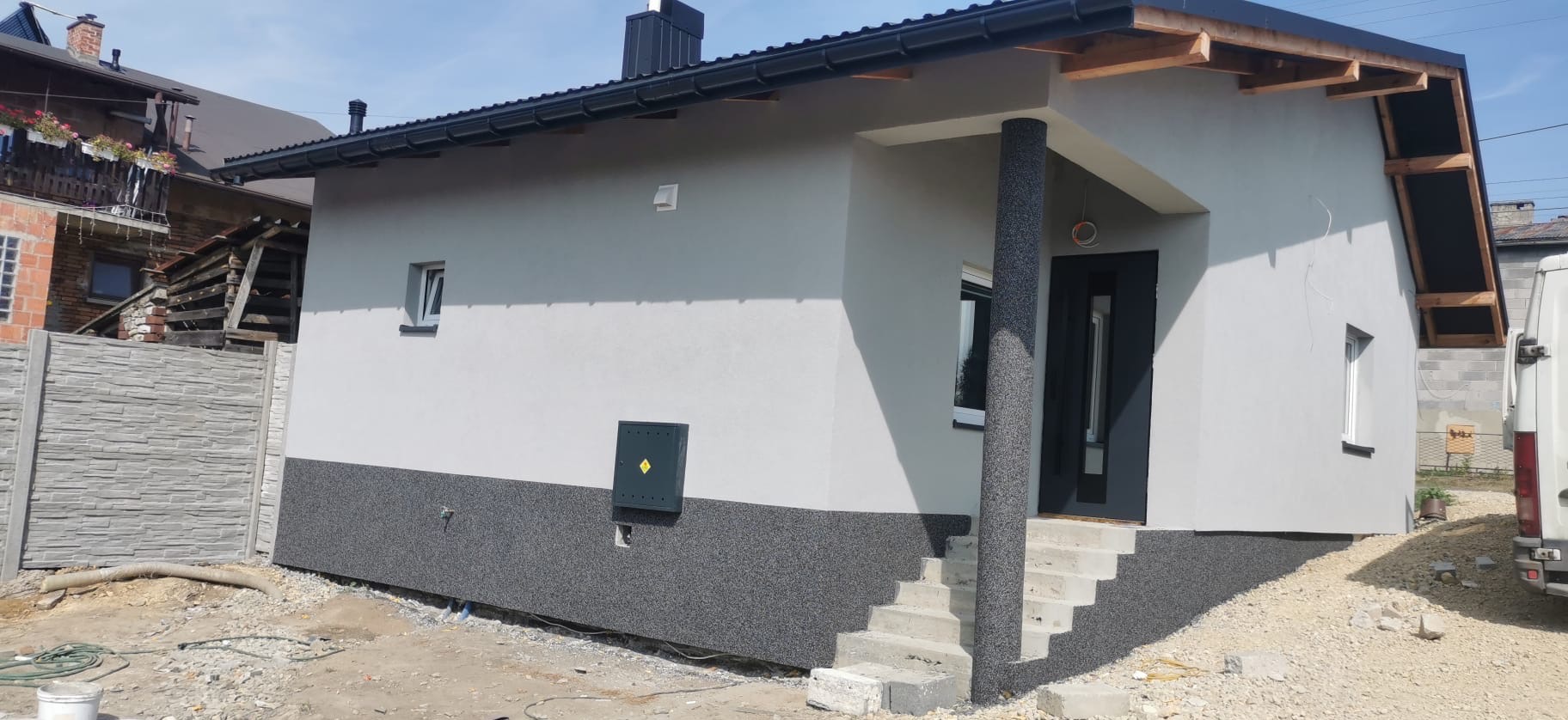 Otynkowany, parterowy dom z czarną stolarką okienną i drzwiową, wykończony szarym tynkiem mozaikowym na cokole, widoczne schody wejściowe z betonowych bloczków i fragment instalacji elektrycznej...