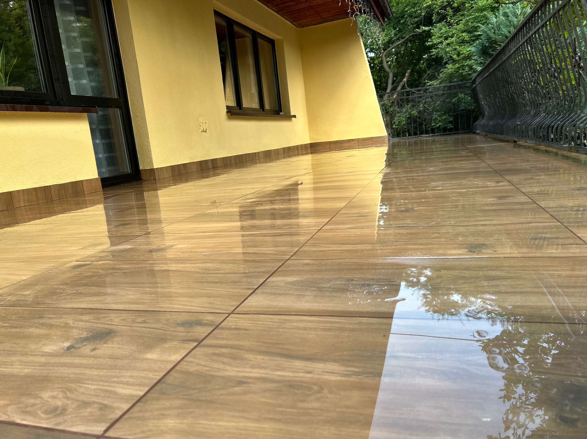 Błyszcząca, brązowa terakota na balkonie domu jednorodzinnego po deszczu, odbijająca w kałużach zieleń drzew i niebo, z widoczną żółtą elewacją i czarnymi ramami okiennymi.