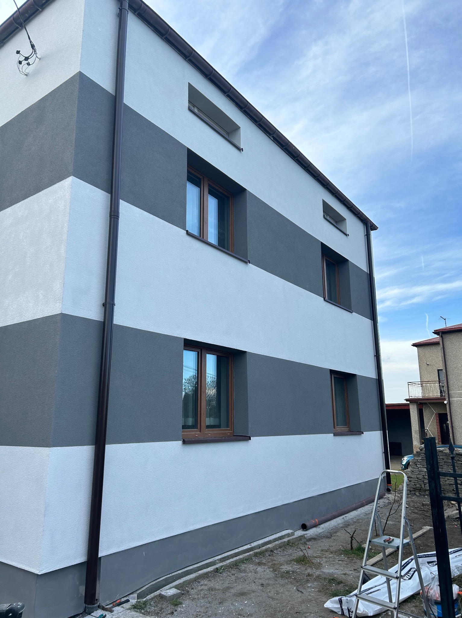 Dwukondygnacyjny dom z ocieploną elewacją w poziome pasy szarości i bieli, z brązowymi oknami i rynnami, widok z dołu na tle błękitnego nieba, drabina oparta o ścianę.