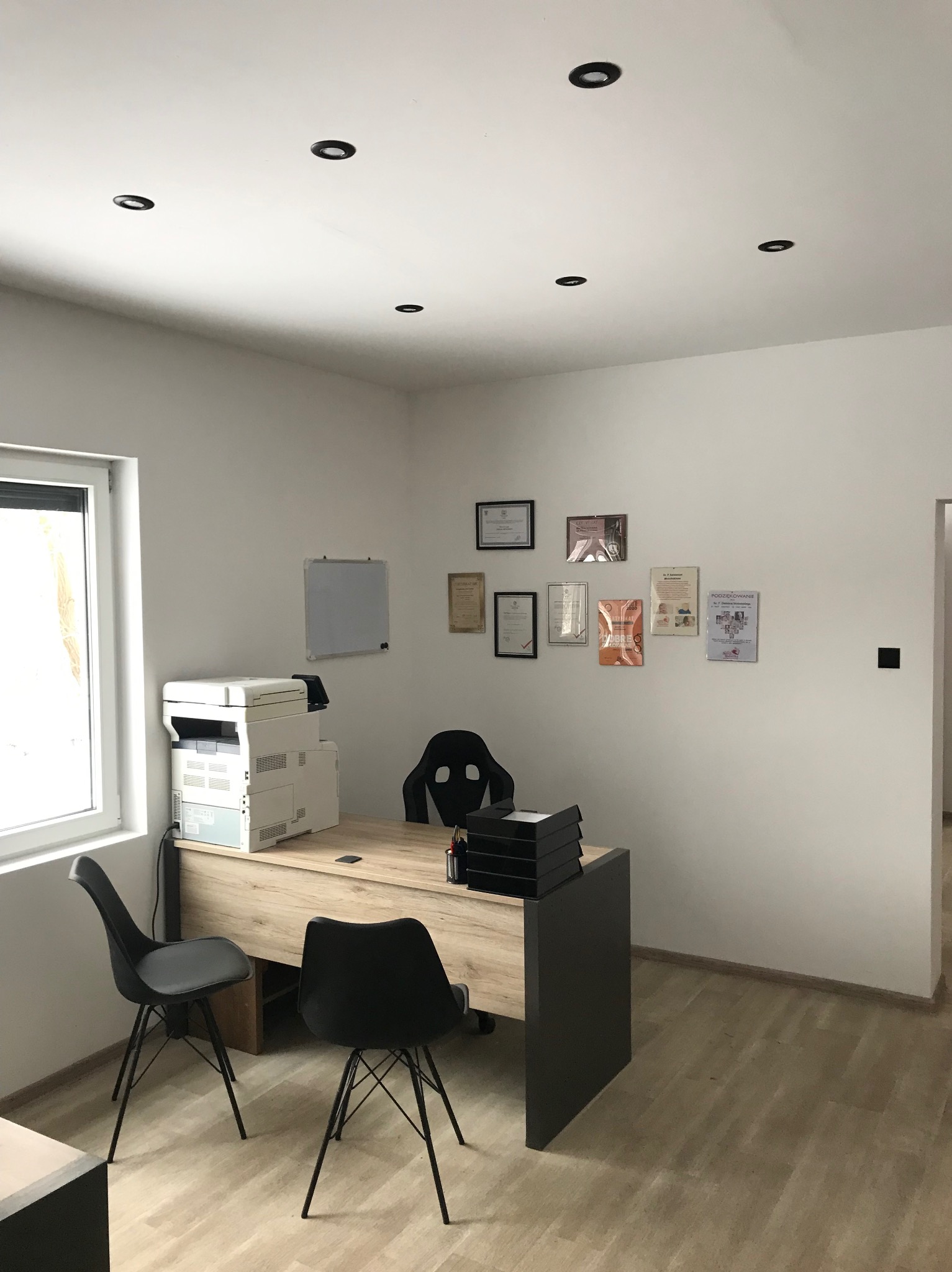 Jasne biuro z drewnianym biurkiem, czarnymi krzesłami i ścianą z certyfikatami, oświetlone punktowymi lampami sufitowymi.