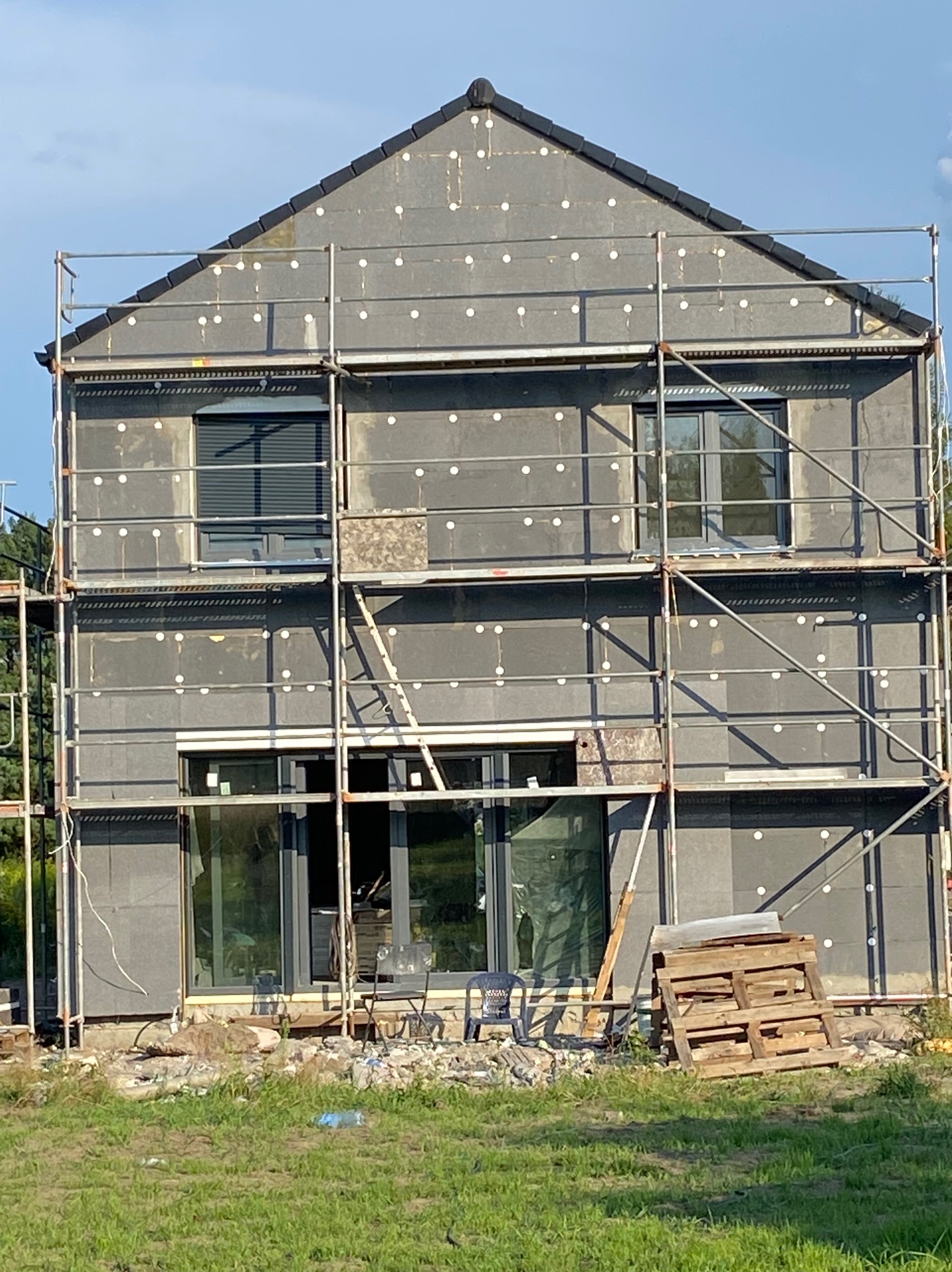 Dom w trakcie ocieplania styropianem grafitowym, widoczne rusztowania, okna z roletami zewnętrznymi, paleta drewniana leżąca na trawie.