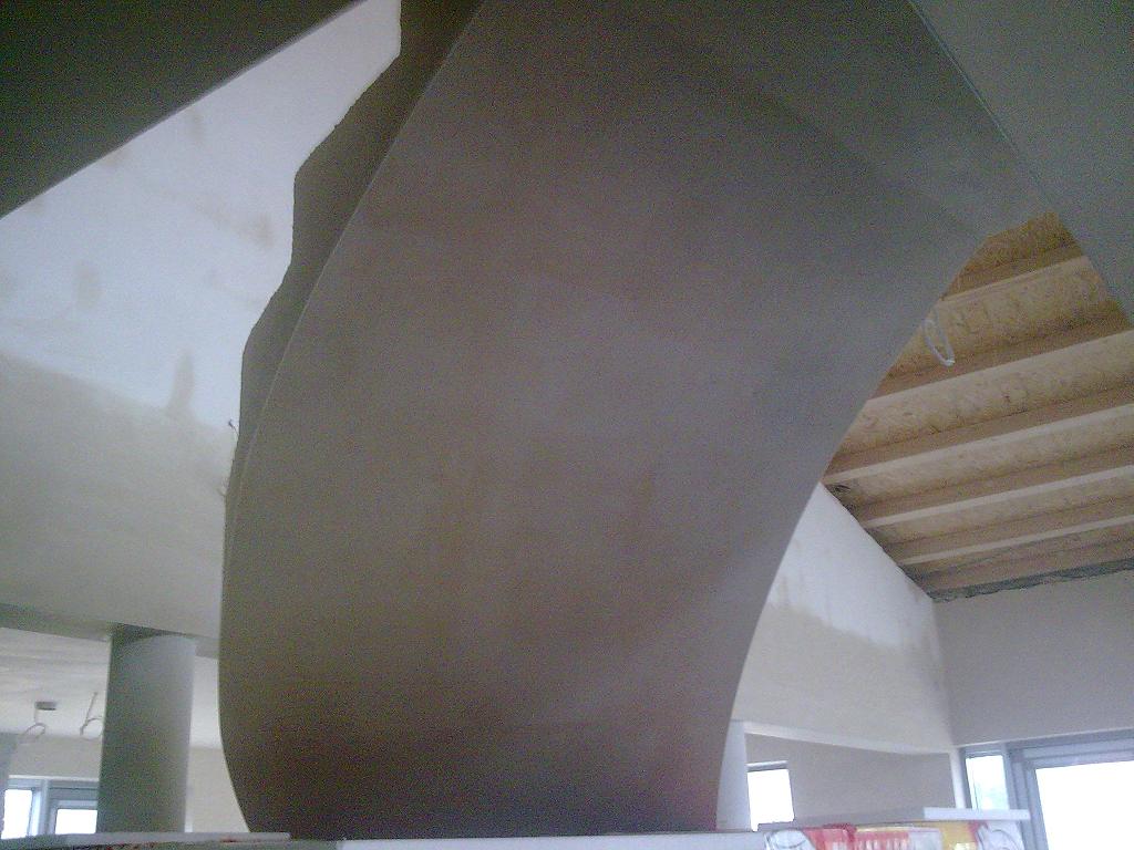 Niedokończone wnętrze z surowymi tynkami maszynowymi na zaokrąglonej ścianie, widoczne drewniane belki stropowe i kolumna nośna.