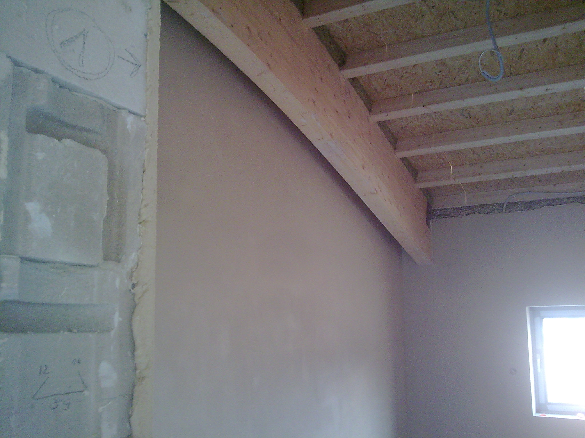 Surowy stan wnętrza z nałożonymi tynkami maszynowymi, widoczna drewniana belka stropowa, fragment ściany z oznaczeniami i okno w tle.