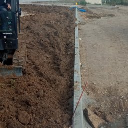 DAWID WILIŃSKI - Mini koparka Yanmar i wykopany rów w ziemi, widoczne krawężniki i budynek przemysłowy w tle.