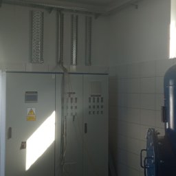 DAWID WILIŃSKI - Wnętrze pomieszczenia z szafami sterowniczymi, metalowymi korytkami kablowymi pod sufitem i dużym niebieskim zbiornikiem ciśnieniowym, widok przez otwarte drzwi na zewnątrz.