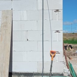DAWID WILIŃSKI - Surowy mur z betonu komórkowego z pionowo poprowadzoną rurką instalacyjną, widoczna łopata i fragment izolacji fundamentu, niezagospodarowany teren budowy.