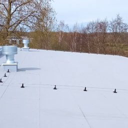 DAWID WILIŃSKI - Widok z góry na płaski, jasny dach z zainstalowanymi uchwytami i przewodem odgromowym, wentylatory dachowe w tle, drzewa bez liści w oddali.