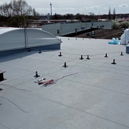 DAWID WILIŃSKI - Szary dach z zamontowanym systemem odgromowym, widoczne uchwyty, linki i narzędzia, w tle budynki przemysłowe i pochmurne niebo.