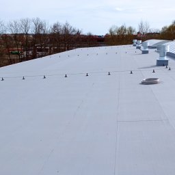 DAWID WILIŃSKI - Biały dach przemysłowy z systemem odgromowym i wentylacją, widok z góry na tle drzew i zabudowań miejskich.