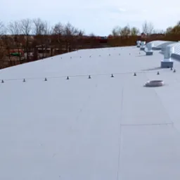 Biały dach przemysłowy z systemem odgromowym i wentylacją, widok z góry na tle drzew i zabudowań miejskich.