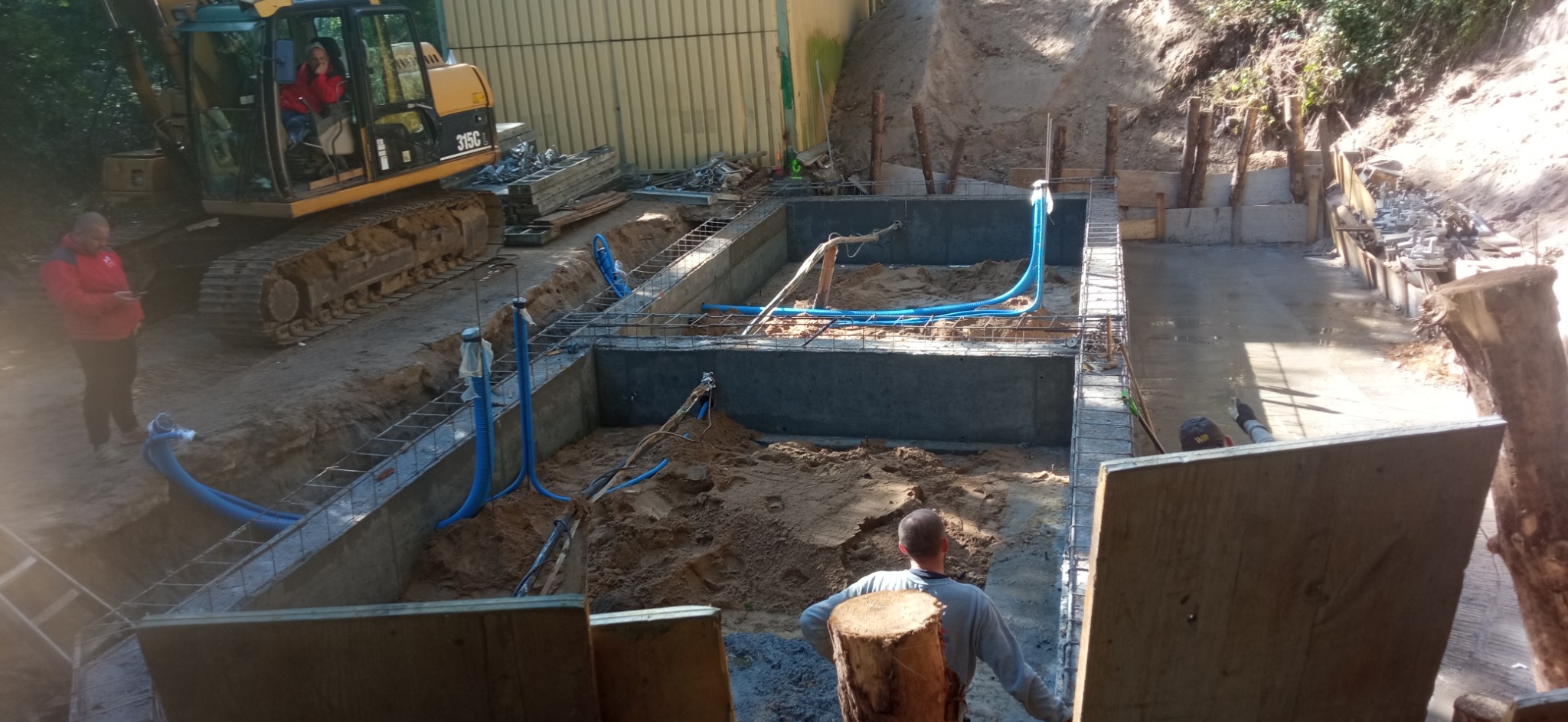 Budowa fundamentów pod basen w Koszalinie z widocznymi niebieskimi rurami instalacji elektrycznej oraz koparką i pracownikami na budowie.