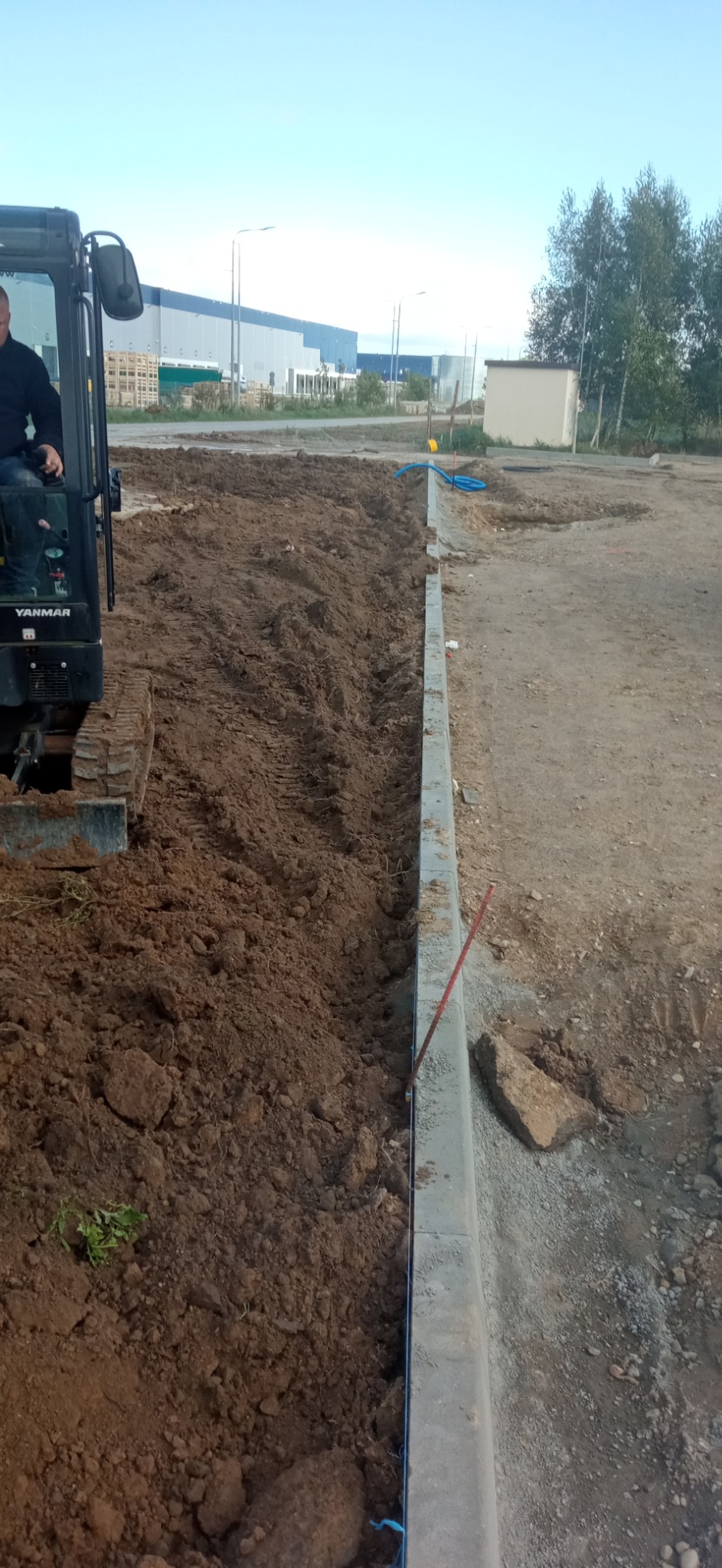 Mini koparka Yanmar i wykopany rów w ziemi, widoczne krawężniki i budynek przemysłowy w tle.
