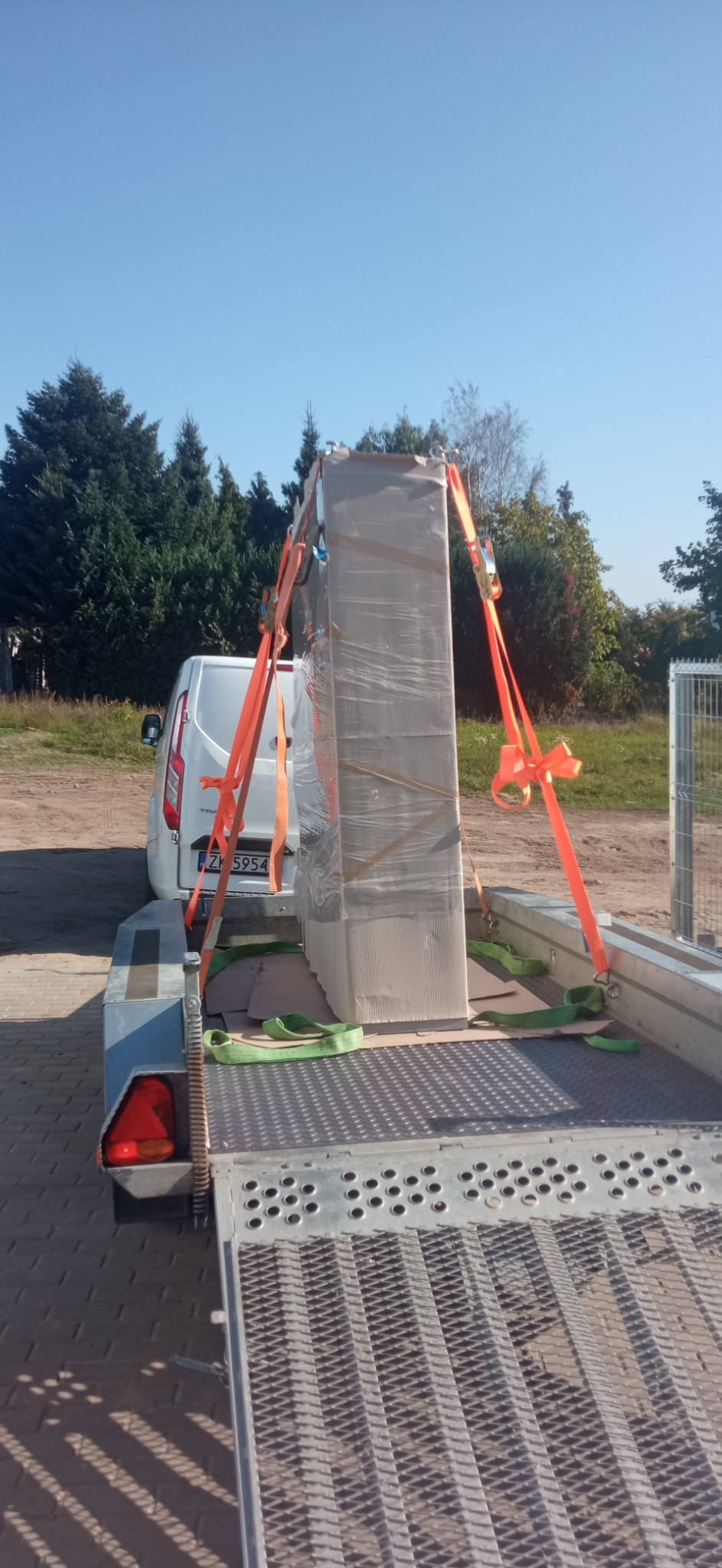 Duża, owinięta folią szafa elektryczna zabezpieczona pomarańczowymi pasami transportowymi na przyczepie samochodowej, widoczny biały samochód dostawczy i błękitne niebo.