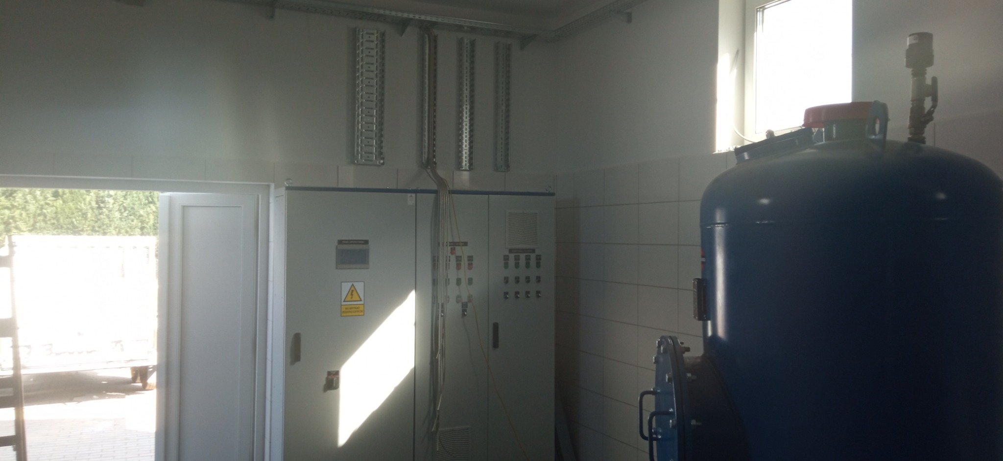 Wnętrze pomieszczenia z szafami sterowniczymi, metalowymi korytkami kablowymi pod sufitem i dużym niebieskim zbiornikiem ciśnieniowym, widok przez otwarte drzwi na zewnątrz.