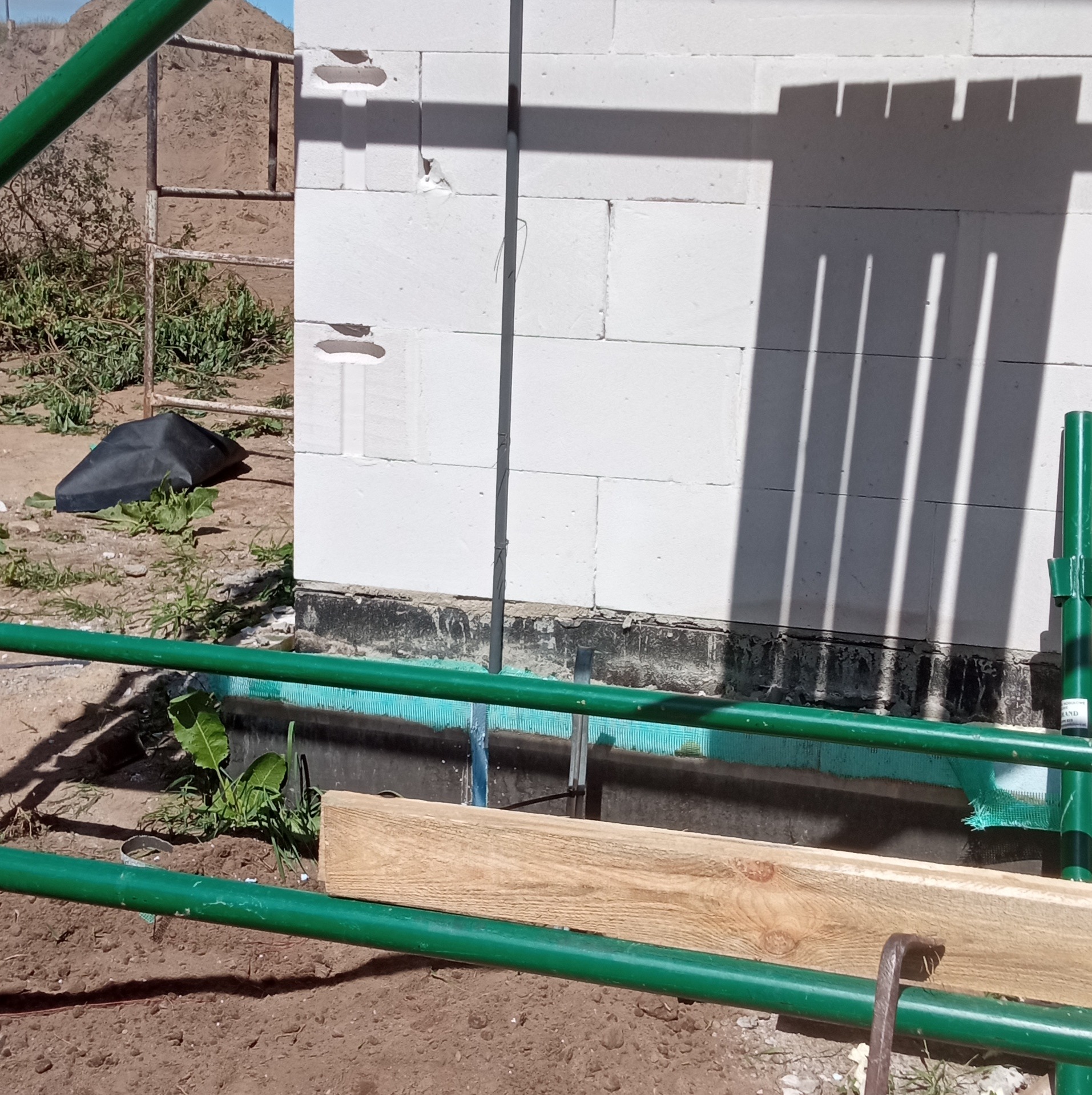 Wznoszenie ściany z białych bloczków, widoczna hydroizolacja fundamentów i fragment rusztowania.