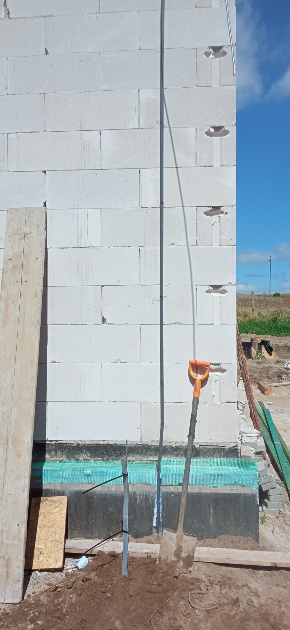 Surowy mur z betonu komórkowego z pionowo poprowadzoną rurką instalacyjną, widoczna łopata i fragment izolacji fundamentu, niezagospodarowany teren budowy.