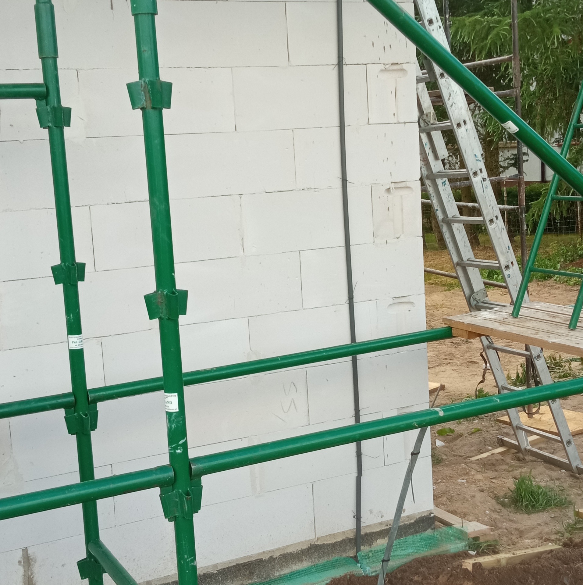 Ściana budynku z betonu komórkowego w trakcie budowy, widoczne zielone rusztowanie i drabina przystawna, przy ścianie pionowy kabel elektryczny.
