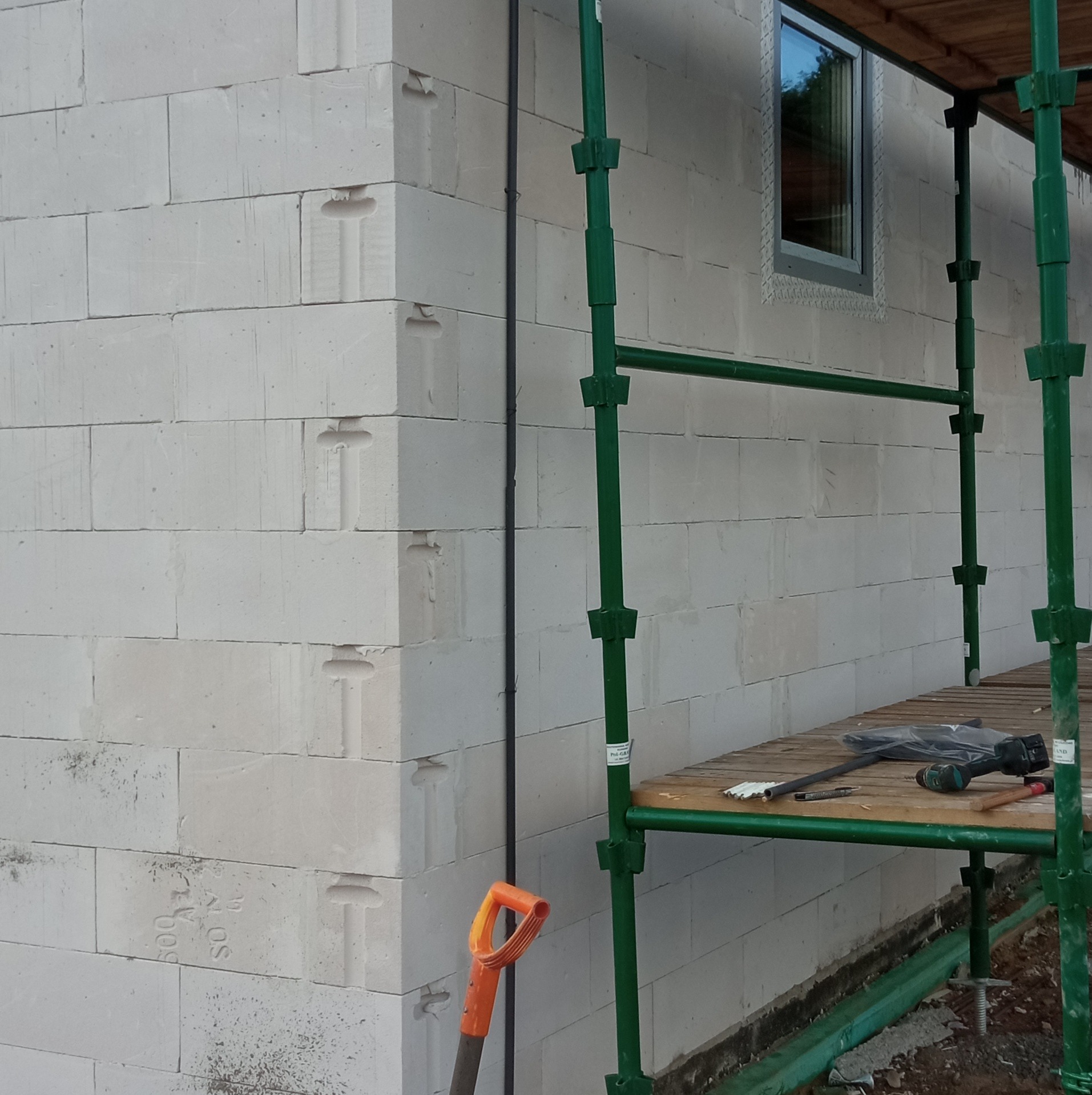 Ściana budynku z betonu komórkowego w trakcie budowy, widoczny kabel elektryczny przymocowany do ściany, rusztowanie, okno i narzędzia na podeście rusztowania.