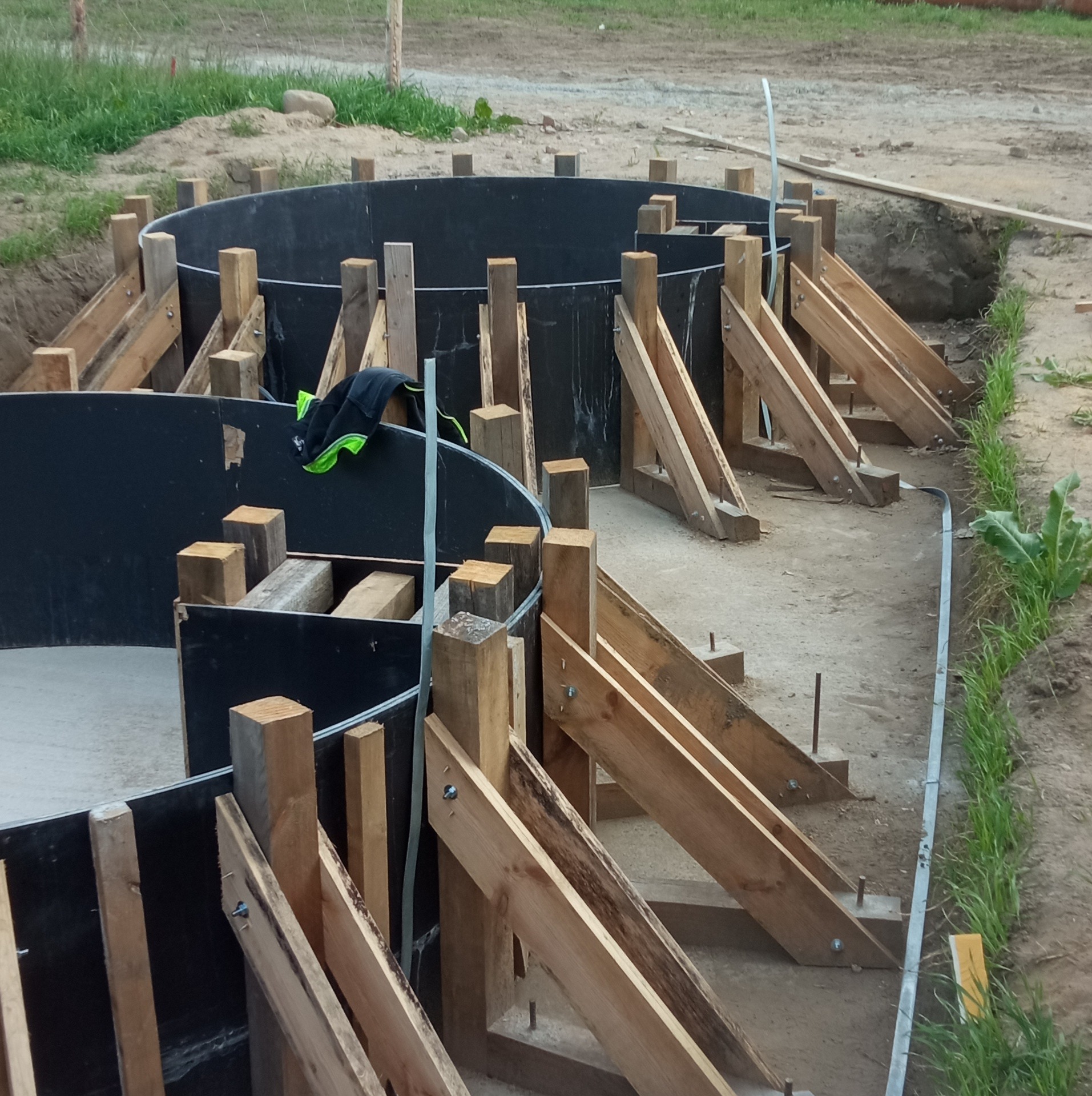 Budowa fundamentów pod okrągłe zbiorniki: widoczne czarne szalunki z drewnianymi podporami na placu budowy, z ziemią i trawą w tle.
