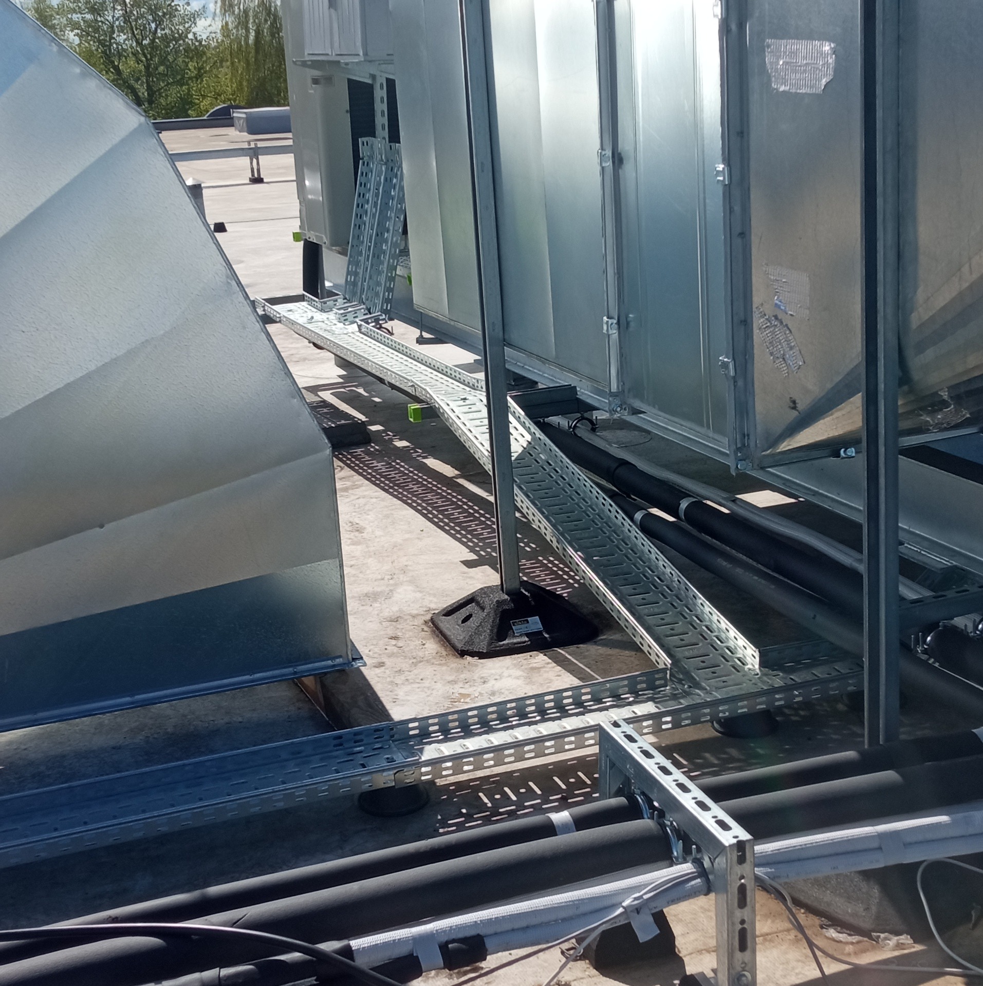 Instalacja na dachu z metalowymi korytkami kablowymi, rurami osłonowymi i urządzeniami klimatyzacyjnymi, widok z góry w słoneczny dzień.