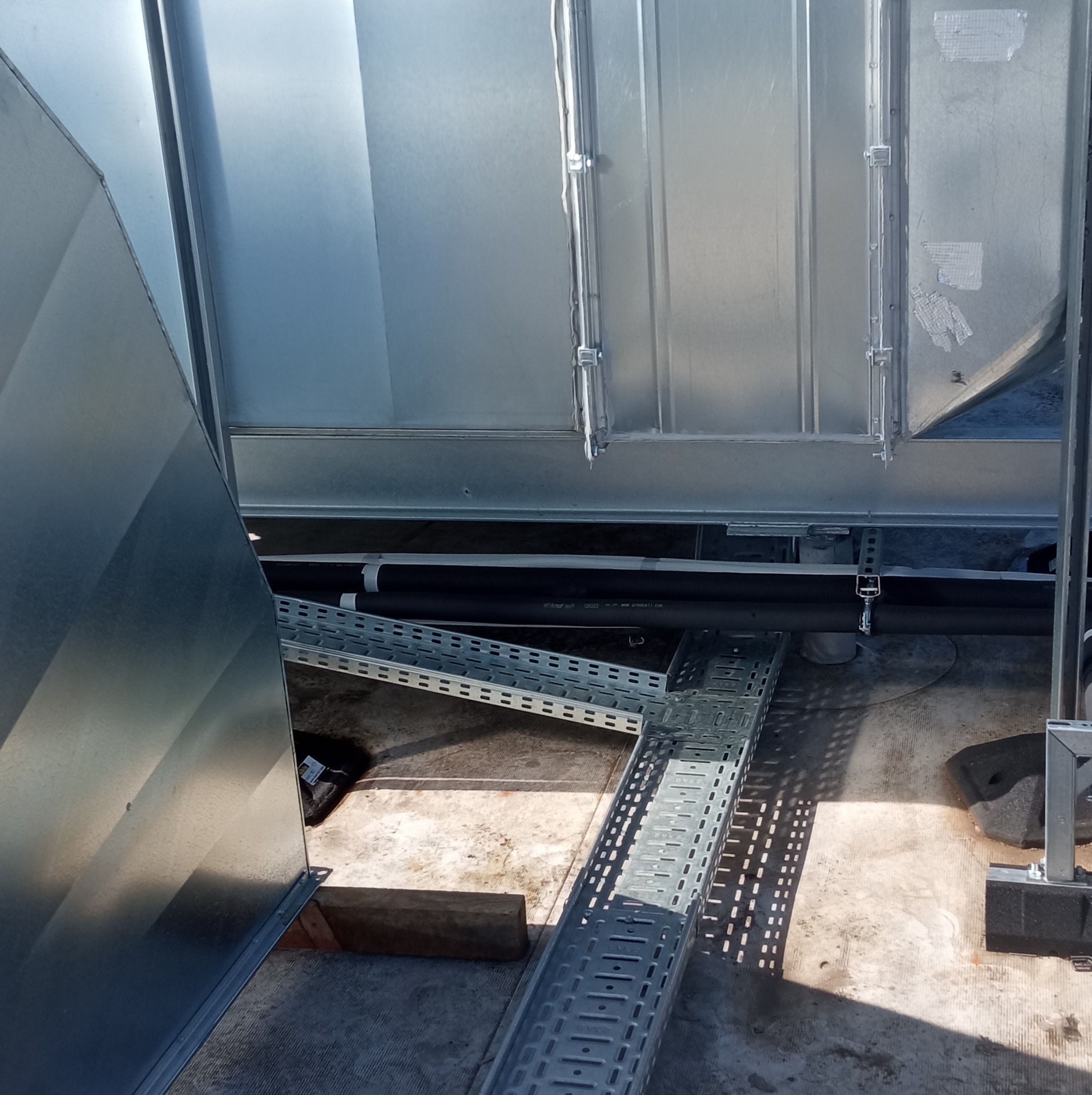 Widok z góry na metalowe kanały kablowe i rury w izolacji, zamocowane do konstrukcji budynku, z widocznymi elementami wentylacji.