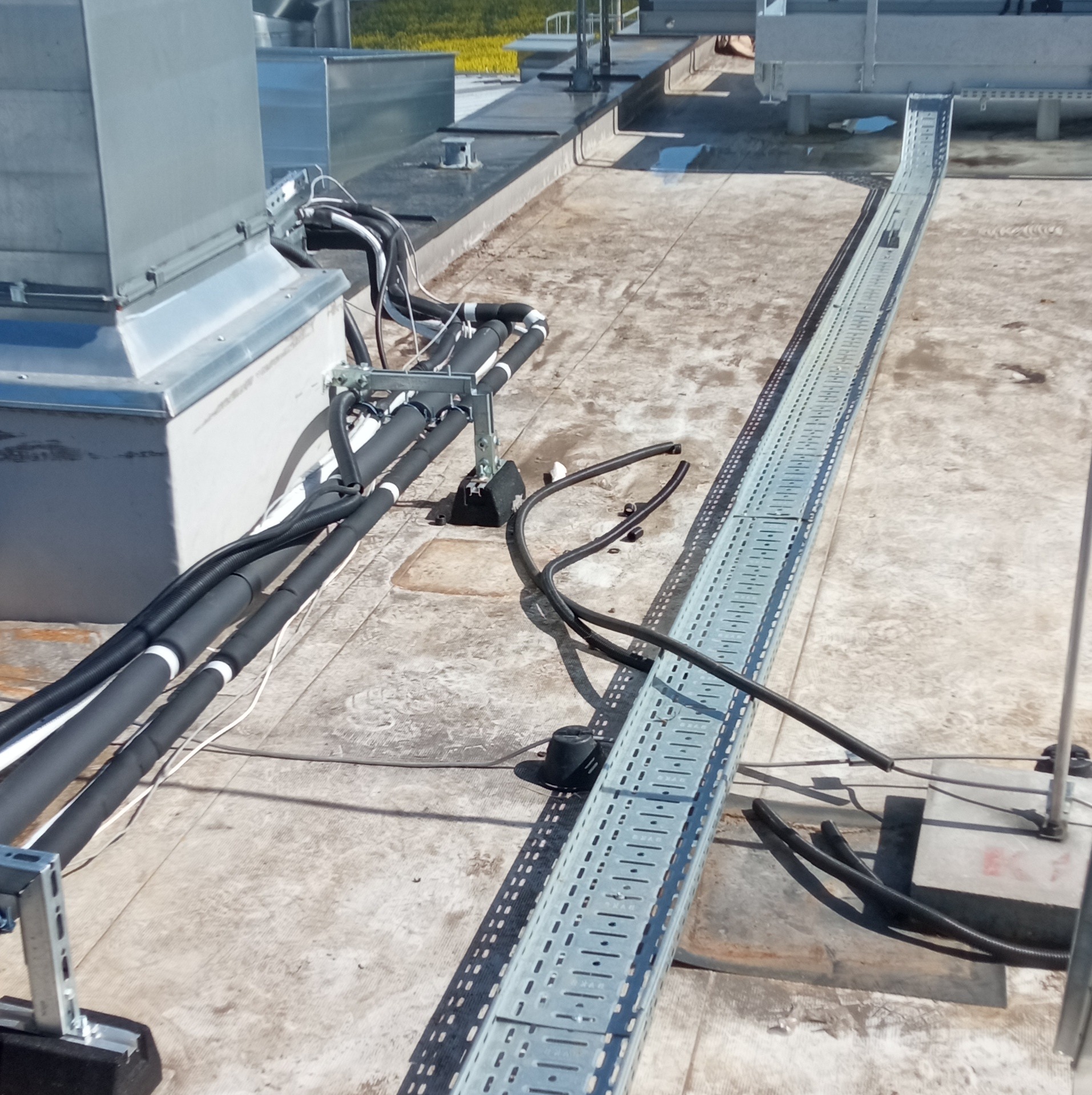 Instalacja kabli w korycie na dachu budynku, widoczne kable w izolacji oraz elementy mocujące, na dalszym planie urządzenia techniczne.