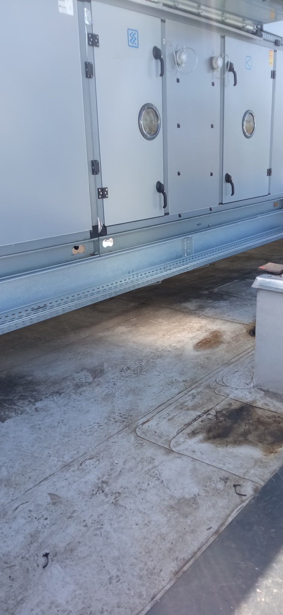 Szara centrala wentylacyjna z czarnymi uchwytami i okrągłymi okienkami inspekcyjnymi, zamontowana na metalowej konstrukcji na dachu pokrytym papą.