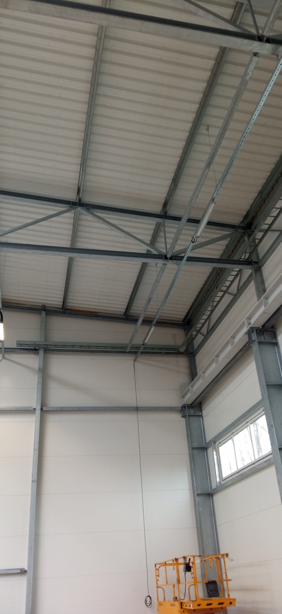Wnętrze hali produkcyjnej w trakcie budowy, widoczna konstrukcja stalowa dachu, ściany z paneli oraz podnośnik koszowy w dolnym rogu.