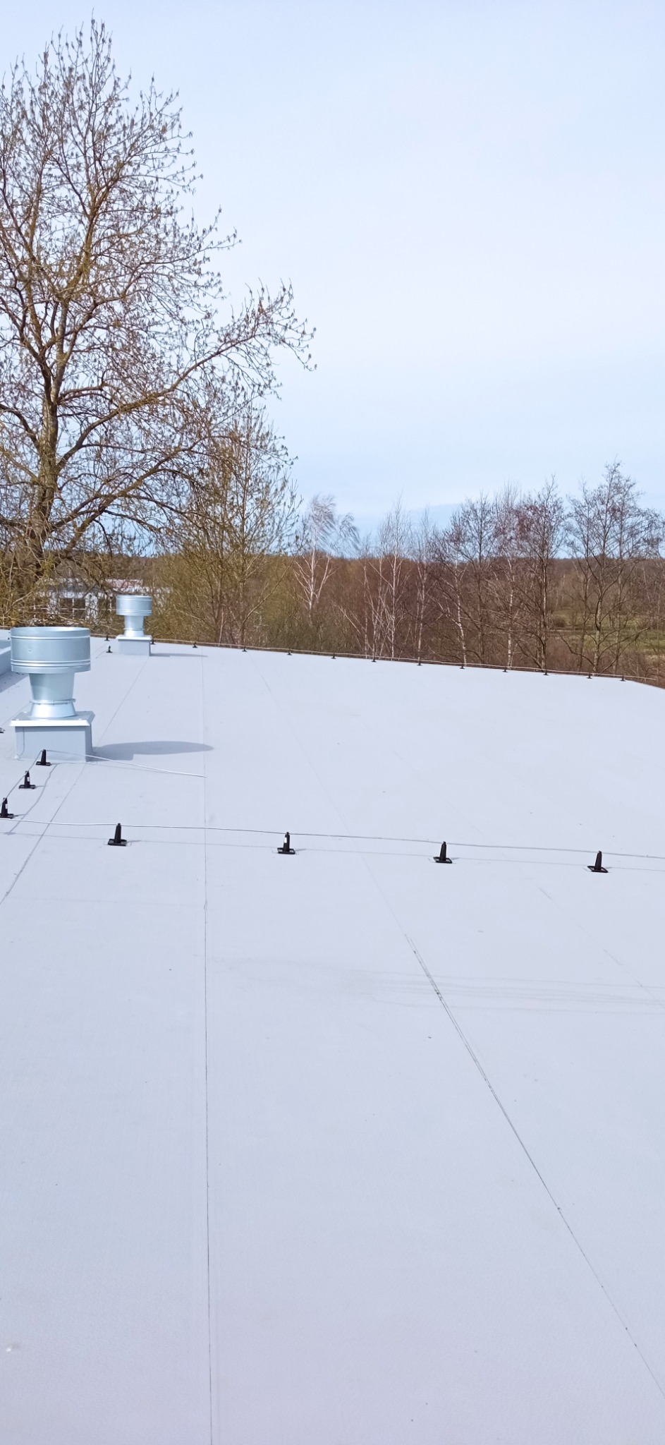 Widok z góry na płaski, jasny dach z zainstalowanymi uchwytami i przewodem odgromowym, wentylatory dachowe w tle, drzewa bez liści w oddali.