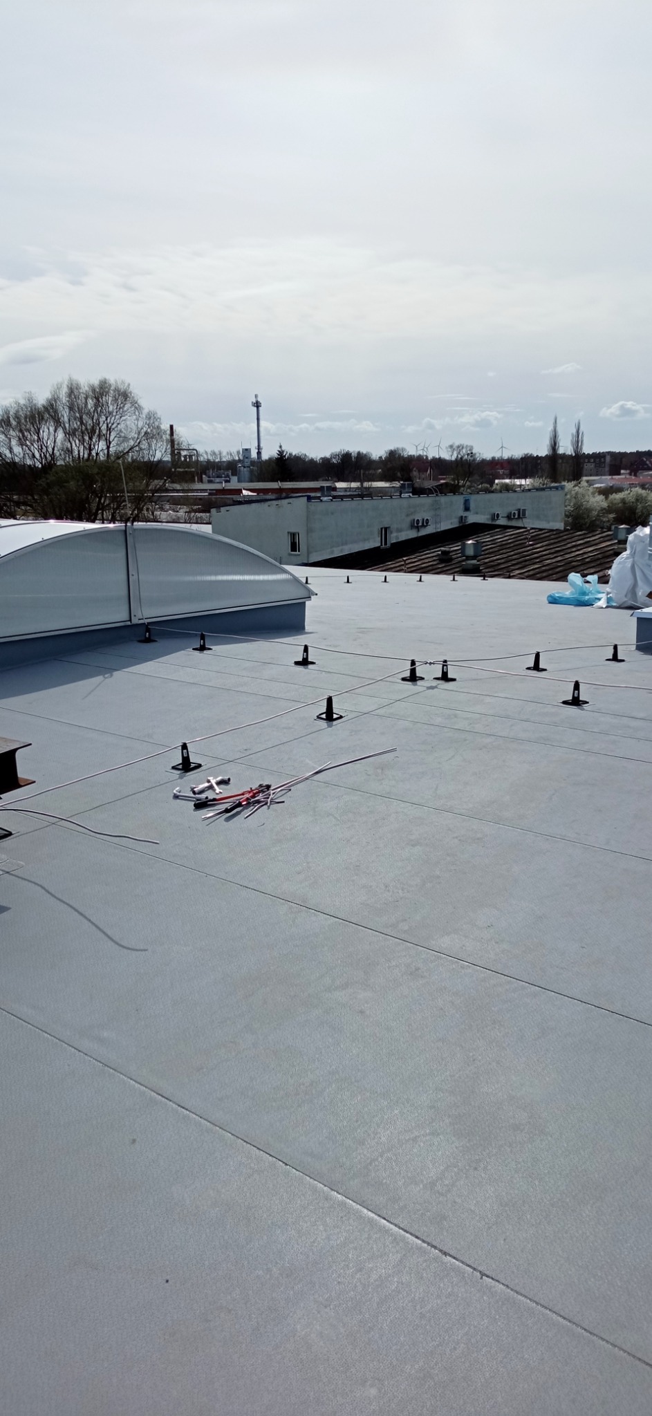 Szary dach z zamontowanym systemem odgromowym, widoczne uchwyty, linki i narzędzia, w tle budynki przemysłowe i pochmurne niebo.