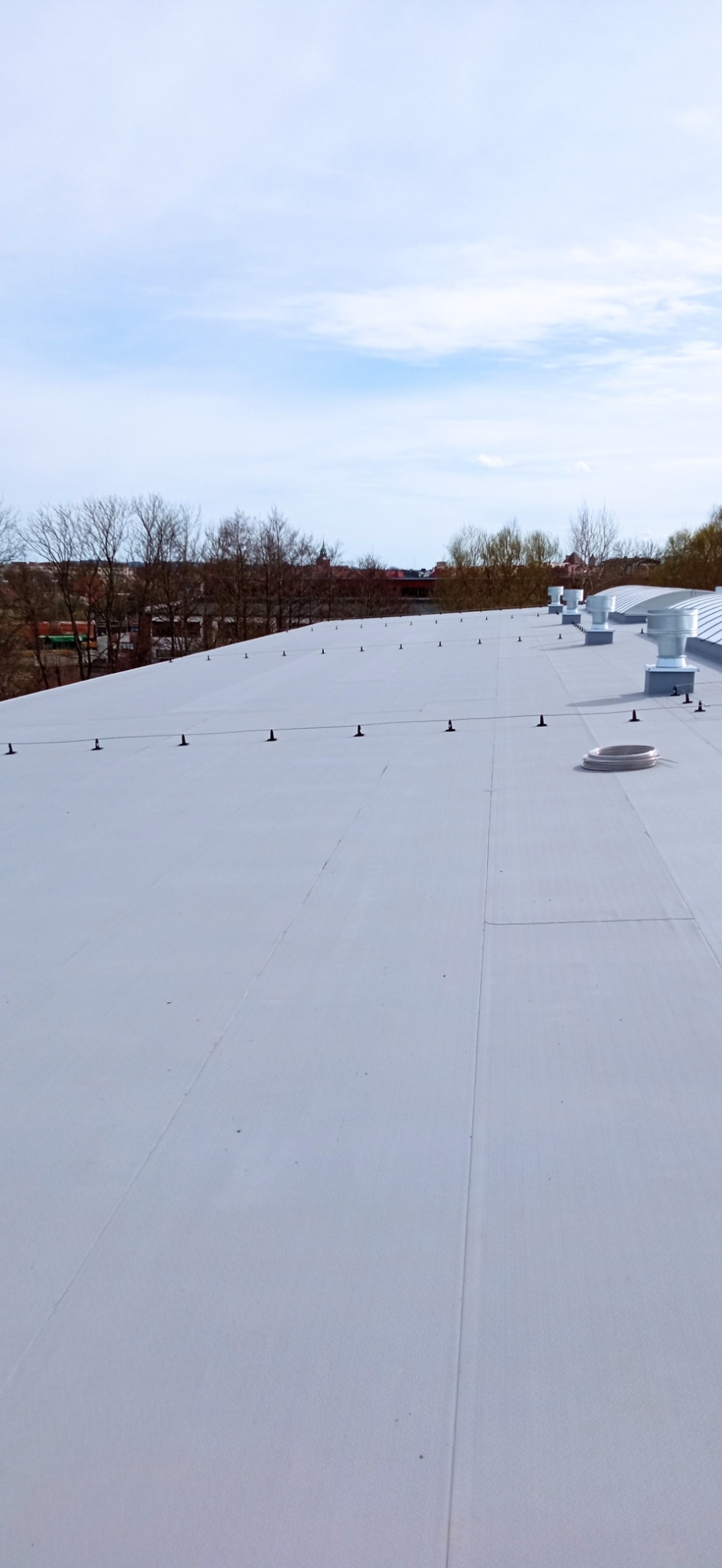 Biały dach przemysłowy z systemem odgromowym i wentylacją, widok z góry na tle drzew i zabudowań miejskich.