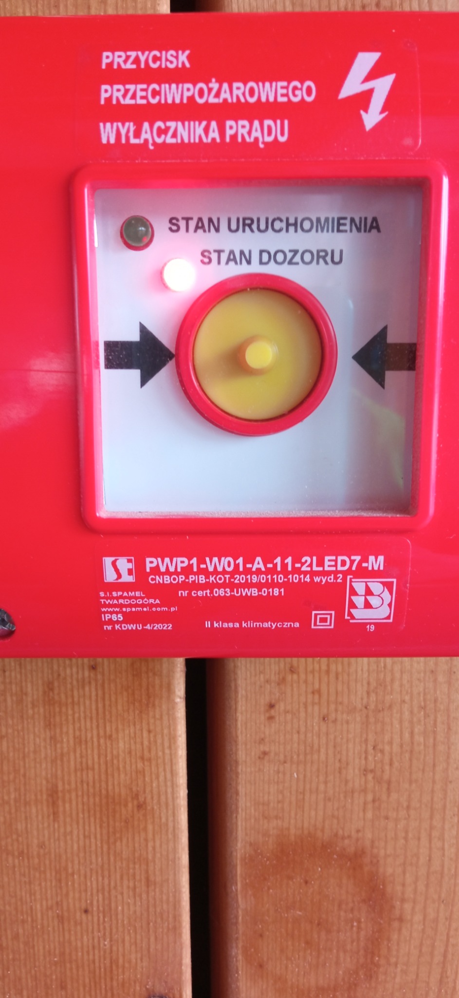 Czerwony przycisk przeciwpożarowy wyłącznika prądu z żółtym elementem centralnym, strzałkami i diodami stanu uruchomienia i dozoru, zamontowany na drewnianej ścianie.