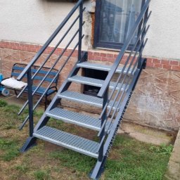 Spaw Serwis Patryk Kubiak - Zewnętrzne metalowe schody kratowe, pomalowane na szaro, prowadzące do piwnicy budynku z ceglaną podmurówką i oknem w czarnej ramie. Widoczny fragment ogrodu z trawą.