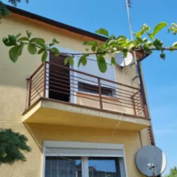 Balkon z brązową, spawaną balustradą w budynku o żółtej elewacji, widok z dołu, częściowo zasłonięty gałęzią z zielonymi owocami.